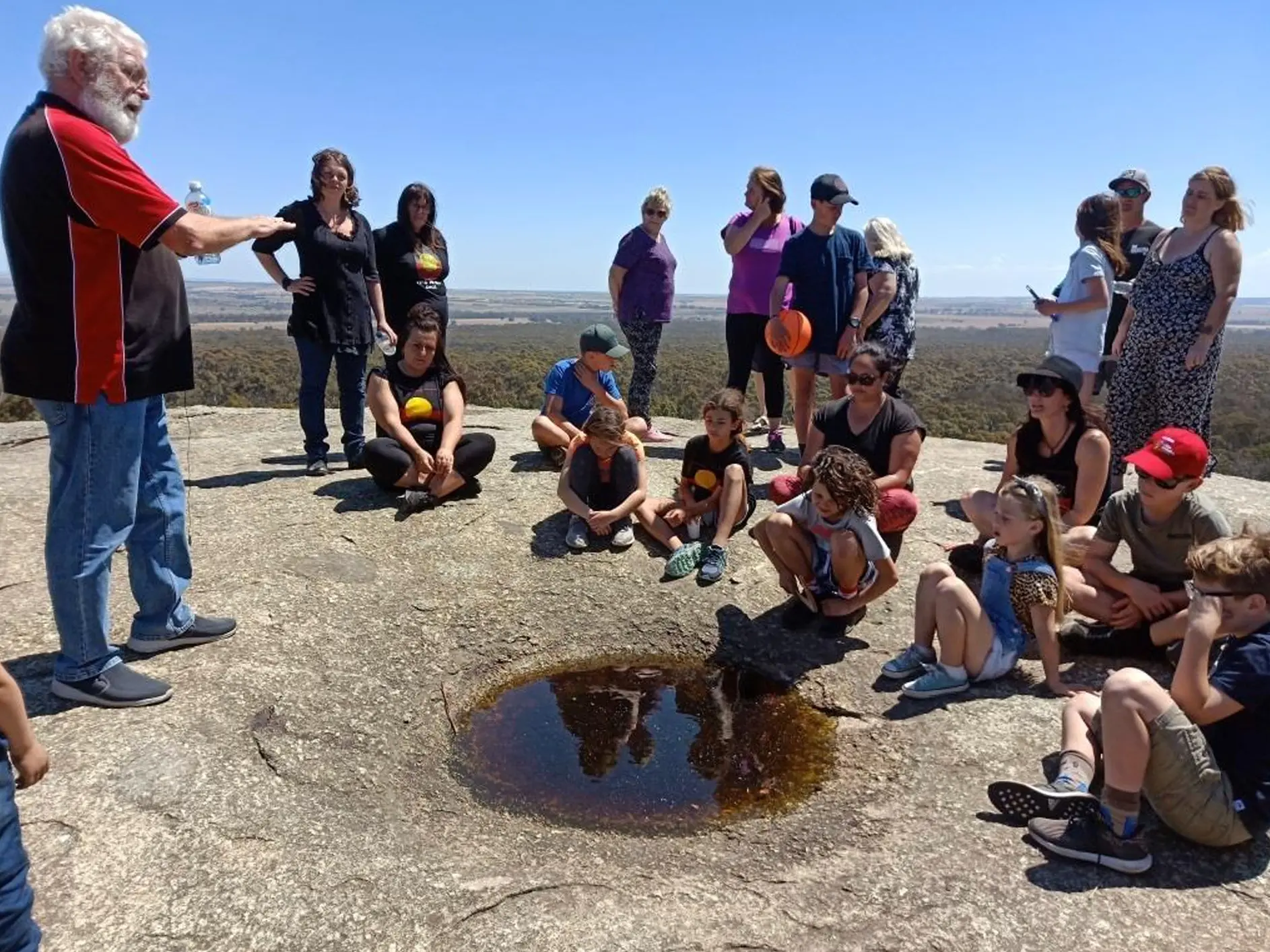 Uncle Bryon sharing knowledge with a group gathered around a rock waterhole at the You Yangs